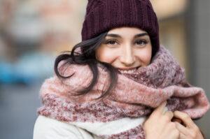 Eine junge, glückliche Frau, die den Winter mit Schal und Mütze genießt und lächelnd in die Kamera schaut.